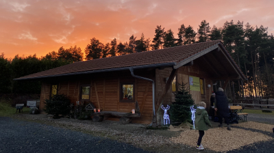 Holzhütte im Christbaumdorf Klug, Treffpunkt für Firmenweihnachtsfeier mit Christbaum selbst sägen Holzhütte bei Sonnenuntergang vor Nadelbäumen, Menschen stehen davor und bereiten eine Firmenweihnachtsfeier vor bei der Christbäume selbst gesägt werden