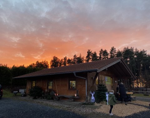 Holzhütte im Christbaumdorf Klug, Treffpunkt für Firmenweihnachtsfeier mit Christbaum selbst sägen Holzhütte bei Sonnenuntergang vor Nadelbäumen, Menschen stehen davor und bereiten eine Firmenweihnachtsfeier vor bei der Christbäume selbst gesägt werden