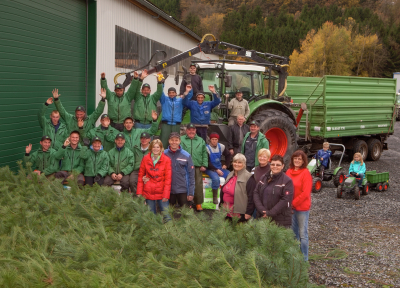 Gruppenfoto von Männern und Frauen in grüner Arbeitskleidung vor einer Halle mit Traktor und aufgestapeltem Tannengrün