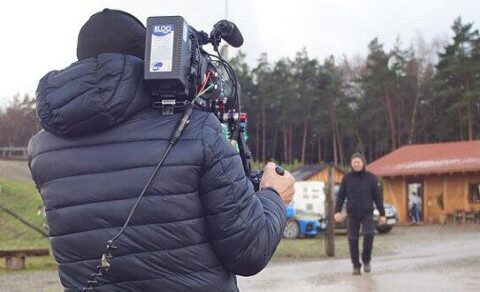 Uwe Klug auf dem Weg zur Plantage – gefilmt vom ZDF Kameramann filmt eine Person beim Gehen über einen Hof, Weihnachtsbäume und Hütten im Hintergrund