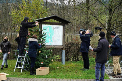 Fernsehleute filmen beim Aufstellen und Schmücken eines Weihnachtsbaumes an einer Informationstafel