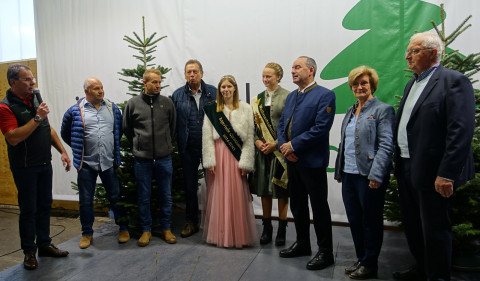 Offizielle Inthronisierung der Christbaumkönigin auf der Weihnachtsbaumbörse Elf Personen stehen auf einer Bühne vor einem Banner mit Tannenbaummotiv, eine Person spricht in ein Mikrofon